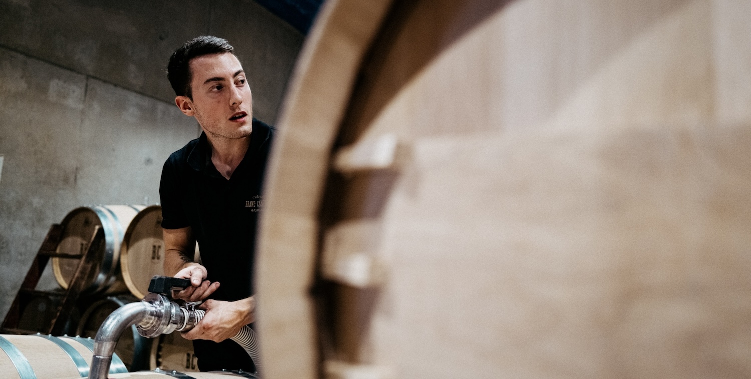 A winery worker at Chateau Brane Cantenac topping up a wooden wine barrel, looking off to the side.